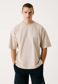 Beige Baumwoll-T-Shirt mit lockerer Passform, kurzen Ärmeln und einem bedruckten Logo auf der Brust. Kombiniert mit dunklen Shorts, die für Freizeitbekleidung entworfen sind.