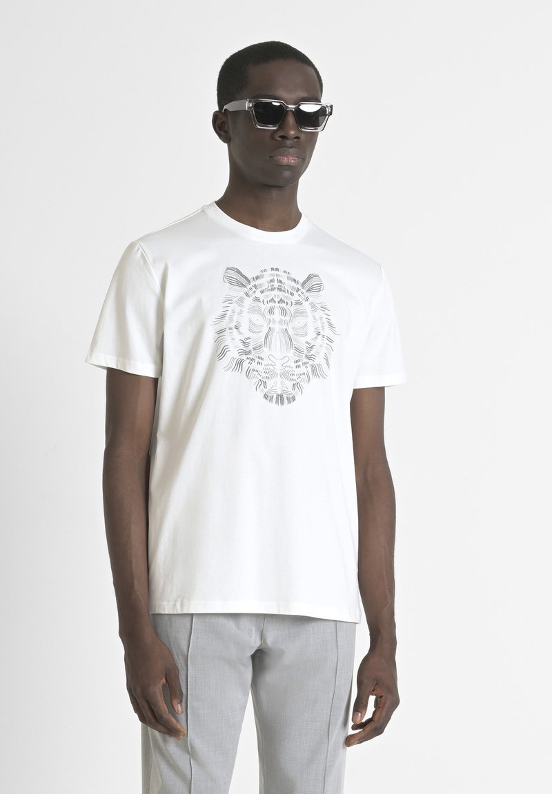 Camiseta de algodón blanca con un gráfico de tigre gris en el frente, combinada con pantalones gris claro y gafas de sol con montura transparente.