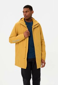 Veste imperméable jaune avec capuche, fermeture à boutons-pression à l'avant et accents réfléchissants. Portée sur une chemise bleue, assortie à un pantalon noir.