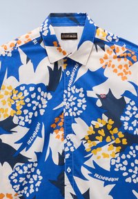 Košile s krátkým rukávem v modré a bílé barvě, s výrazným květinovým vzorem s oranžovými a žlutými akcenty. Knoflíky jsou modré, s detailem loga.