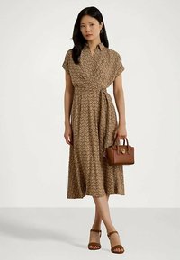 Γυναίκα που φοράει ένα καφέ μίντι φορεματάκι με σχέδιο, κρατώντας μια μικρή καφέ τσάντα χεριού, και φορώντας καφέ πέδιλα με τακούνι.