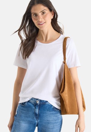 Kvinde iført en hvid t-shirt og blå jeans, bærende på en stor brun ruskindsskuldertaske, smilende med løst mørkt hår.