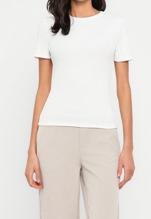 Kvinde iført en almindelig hvid kortærmet tætsiddende T-shirt og lyse beige bukser, stående mod en hvid baggrund.