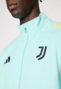 Helehall sinine tõmblukuga jope, millel on must logodetail, kollased ja roosad aktsendid ning kerge, tekstuuriga kangas.