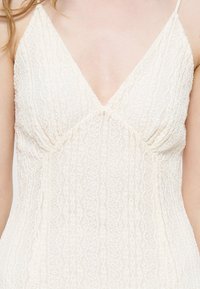 Femme portant une robe blanche en dentelle sans manches avec un décolleté en V profond et un motif texturé, raccourcie aux épaules et au milieu du torse.