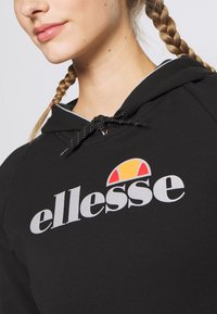 Czarna bluza z kapturem wykonana z miękkiego materiału, z zapięciem na sznurek, z odważnym białym logo "ellesse" i kolorowym akcentem nad nim.