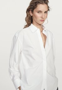Camicia bianca con bottoni, realizzata in cotone, con colletto, maniche lunghe e vestibilità relaxed. Design minimalista senza fantasie o dettagli.