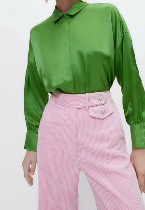 Mujer que lleva una blusa verde brillante de manga larga metida en unos pantalones de pana rosa de talle alto con botones decorativos en la cintura.