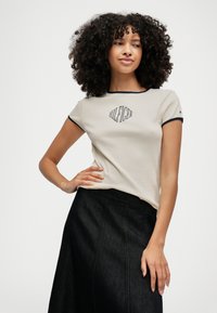 Tricou scurt cu mâneci scurte, de culoare bej deschis, cu bordură bleumarin, având logo-ul "HILFIGER" într-o formă de diamant, poziționat la centru. Asortat cu o fustă din denim închis.