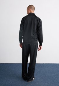 Salopetă neagră dintr-un material neted, cu jachetă cu fermoar și pantaloni cu talie elastică, prezentată din spate pe un fundal alb simplu.