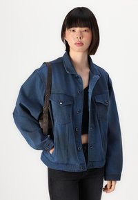 Μπλε τζιν μπουφάν με δύο μπροστινές τσέπες, κλείσιμο με κουμπί και χαλαρή εφαρμογή. Συνοδεύεται από σκούρο καφέ λουρί ώμου.