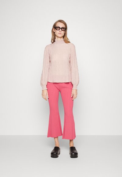 Γυναίκα φορώντας μια υφασμάτινη ανοιχτό ροζ μπλούζα με μακριά μανίκια, ριγωτό ροζ παντελόνι με ανοιχτή γραμμή, μαύρα παπούτσια με πλατφόρμα και μαύρα ορθογώνια γυαλιά, καθώς στέκεται σε γκρι δάπεδο.