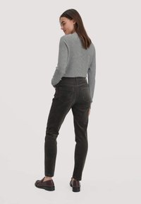 Vrouw die zijwaarts staat, gekleed in een grijze langmouwtop, donkerbruine corduroypantalon en donkerbruine loafers tegen een effen achtergrond.