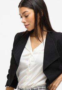 Černá zkrácená bunda s šálovým límcem, nošena přes bílou blůzu s knoflíkovou légou. Model nosí vzorované kalhoty, viděno z boku.