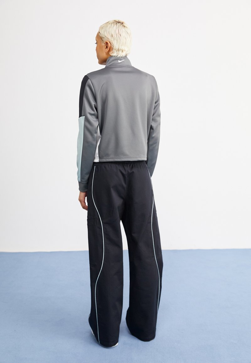 Chaqueta deportiva gris con cuello alto y acentos en azul claro, combinada con pantalones anchos negros con bolsillos laterales y detalles en raya blanca.