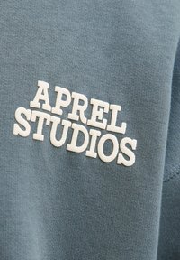 Modrá látka s vyšívaným bílým textem "APREL STUDIOS." Hladká textura a uvolněný střih, který zdůrazňuje prvky ležérního designu.