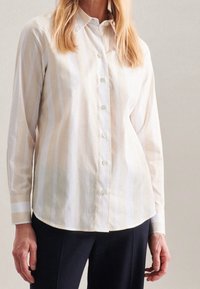 Woman wearing a beige and white vertical striped button-up shirt with black pants against a plain background.