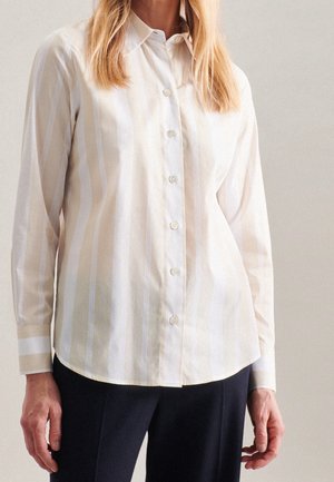 Donna che indossa una camicia a righe verticali beige e bianche con pantaloni neri su uno sfondo semplice.