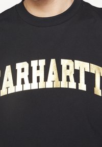 Černé bavlněné tričko s výrazným zlatým foliovým zakřiveným logem "CARHARTT" přes hruď. Klasický kulatý výstřih s hladkou strukturou.