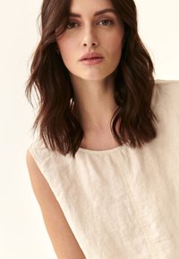 Beige lina tops ar apaļu kakla izgriezumu un bez piedurknēm, ar smalku tekstūru un vertikālu centrālo šuvi. Dabisks, viegls audums.