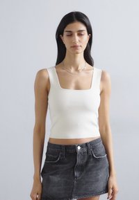 Balta griesta trumpa palaidinė su kvadratine iškirpte, derinama su juoda džinsine sijoną su aukštu liemeniu, turinčia dvi priekinės kišenes.