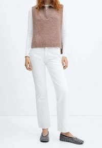 Brauner ärmelloser Strickpullover mit geripptem Saum, getragen über einem weißen Langarmshirt und kombiniert mit weißen Jeans mit geradem Bein und schwarzen Ballerinas.