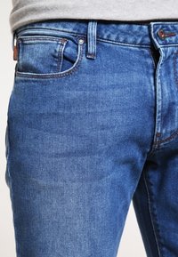 Blue denim jeans with a straight cut, five pockets, and subtle fading, featuring a metal button and zip fly closure.