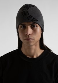 Gorro de punto gris con un patrón de logo negro, usado por un modelo con cabello largo. El modelo lleva una camiseta negra y está frente a un fondo neutral.