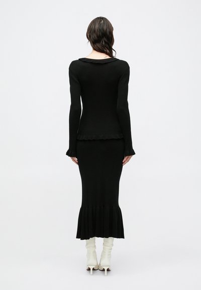 Mustast trikotaažist kleit istuva ülakeha, lehviva ääre ja soonilise tekstuuriga. Pikkadel varrukatel on voltitud mansetid. Kandmisel valgete kontsakingadega.