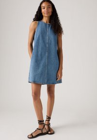 Denim sleeveless dress in light blue with a straight cut, front seam detail, and a hemline above the knee. Paired with black strappy sandals.