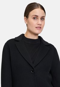Sort uldblandingsjakke med spidse revers, enkeltknaplukning og subtil tekstur. Båret over en sort top med minimalistisk design.