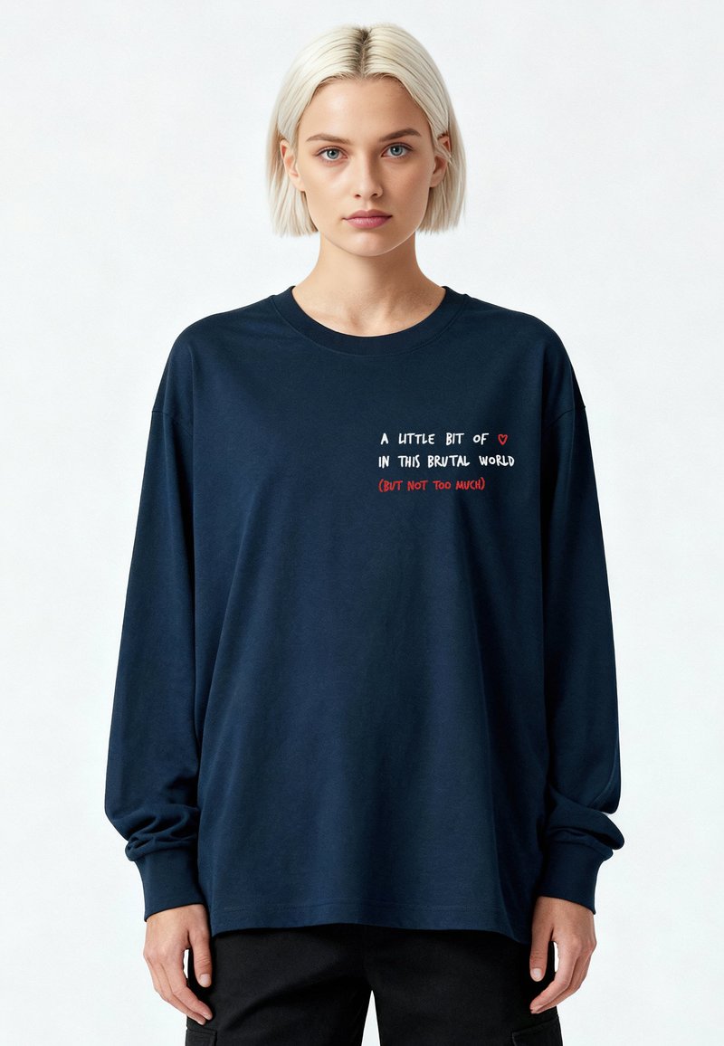Jeune personne aux cheveux blonds courts portant un t-shirt à manches longues bleu marine avec un texte blanc et rouge sur la poitrine.
