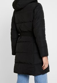 Abrigo de plumas negro con capucha, que presenta una cintura ceñida con cinturón, textura acolchada horizontal y mangas largas.