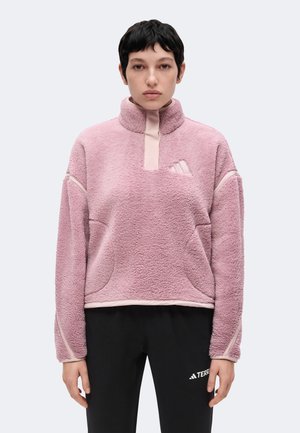 Person iført en lyserød fleece-pullover med høj krave og sorte bukser med hvid logo-tekst på en ensfarvet baggrund.
