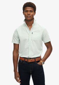 Camicia verde chiaro con maniche corte e colletto abbottonato, tasca sul petto sinistro, abbinata a jeans scuri e una cintura in pelle marrone. Orologio marrone visibile.