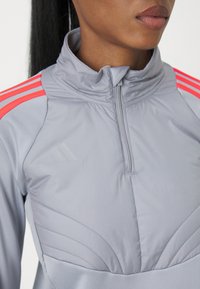 Primo piano di una persona che indossa un top sportivo grigio con zip, con strisce neon rosa sulle spalle e un logo sottile sul petto.