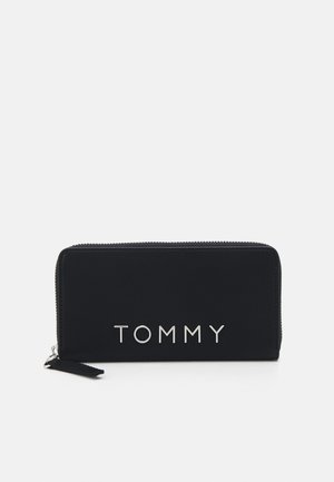 Cartera negra con cremallera hecha de tela suave, que presenta un logo metálico plateado "TOMMY" en la parte frontal, con un tirador de cremallera texturizado.