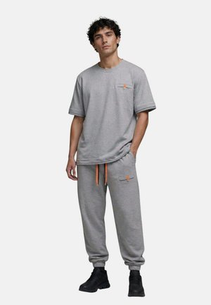Jeune homme portant une chemise grise à manches courtes assortie et un pantalon de jogging avec des cordons orange et des chaussures noires, debout avec une main dans la poche.