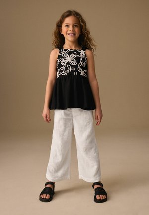 Ragazza sorridente con capelli ricci indossa una canottiera nera con motivi floreali bianchi, pantaloni bianchi larghi e sandali neri, in piedi su uno sfondo beige.