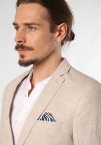 Lichtbeige linnen blazer over een witte henley shirt, met een blauw-wit gestreepte pochet in de borstzak. Zachte textuur.