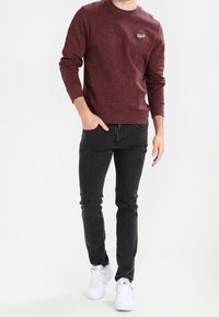 Muž oblečený v vínovém dlouhém svetru, černých slim-fit džínech a bílých teniskách, stojící s jednou rukou v kapse.