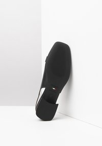 Zapato de tacón alto negro con parte superior de cuero liso, lados abiertos y suela de goma texturizada. Presenta un logo metálico y detalle de la bandera italiana.