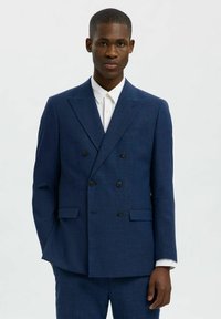 Giacca di suit navy doppiopetto realizzata in tessuto testurizzato, con rever a punta e bottoni neri. Indossata sopra una camicia bianca con colletto.