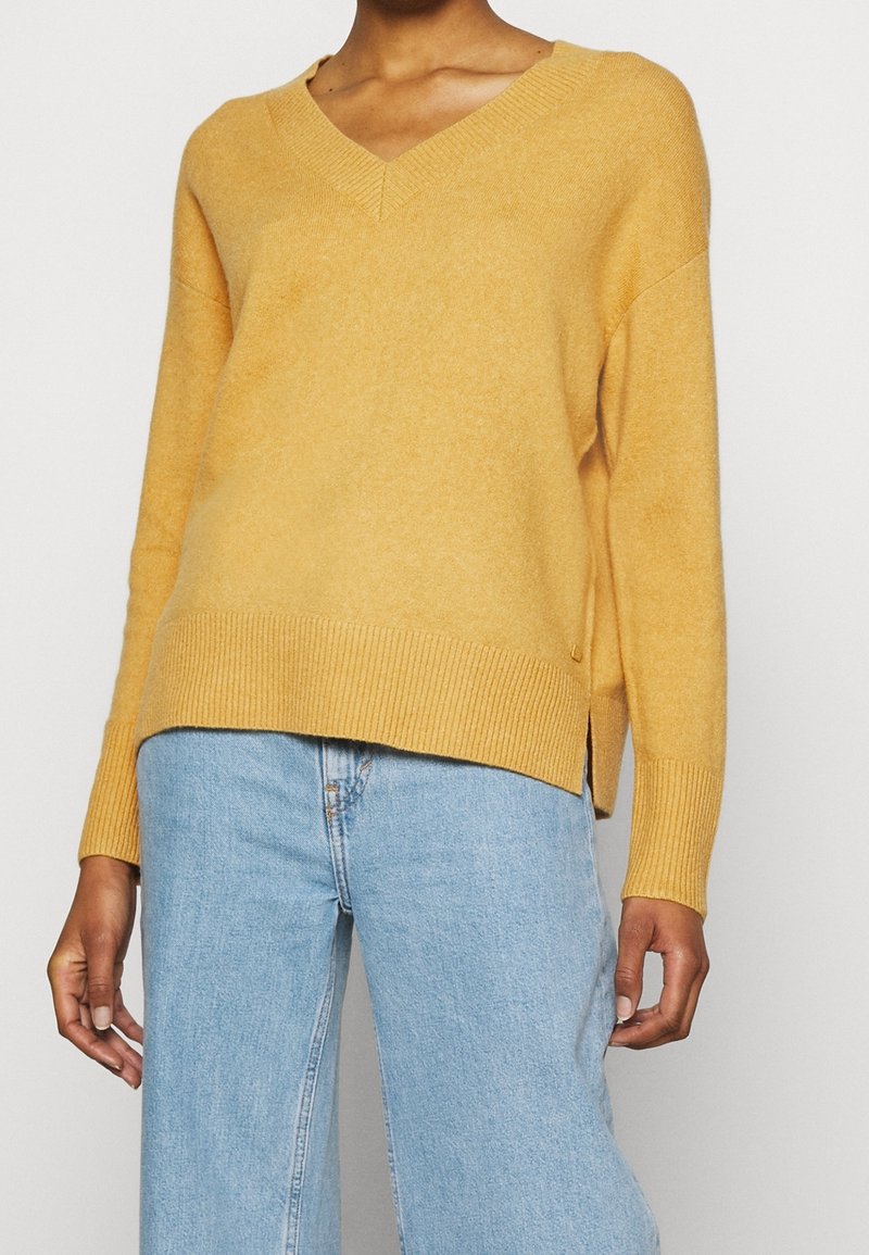 Femme portant un pull jaune moutarde à encolure en V avec des poignets et un ourlet côtelés, assorti à un jean taille haute bleu clair.