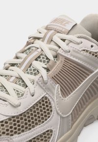 Baskets Nike Vomero 5 beige et blanches avec des panneaux en mesh, des lacets blancs, et un grand swoosh Nike sur le côté.