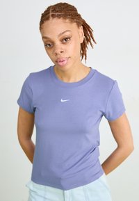 Jauna moteris su rausvais pynimais ir strazdanais, vilkinti šviesiai purpurinę "Nike" marškinėlį ir šviesiai mėlynas džinsus, stovi prieš baltą foną.