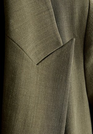 Olivgrüner Stoff mit einer texturierten Oberfläche und eckigen Nahtdetails. Fokus auf das geschichtete Design und kontrastierende Nähtelemente.