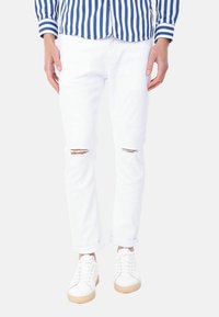 Jeans bianchi strappati con dettagli sfilacciati sulle ginocchia, abbinati a sneaker bianche con suola beige. L'outfit è completato da una camicia a righe blu.