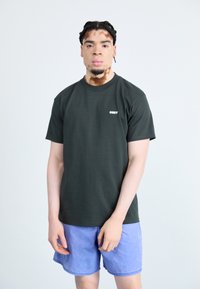 Dark green cotton t-shirt with a crew neck and short sleeves, featuring a white "OBEY" logo on the left side, paired with light blue shorts.