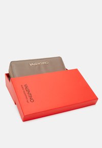 Portofel cu textură maro, cu logo-ul auriu "VALENTINO", parțial vizibil într-o cutie roșie strălucitoare cu text negru.